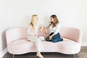 Two women sitting on a pink couch smiling at one another