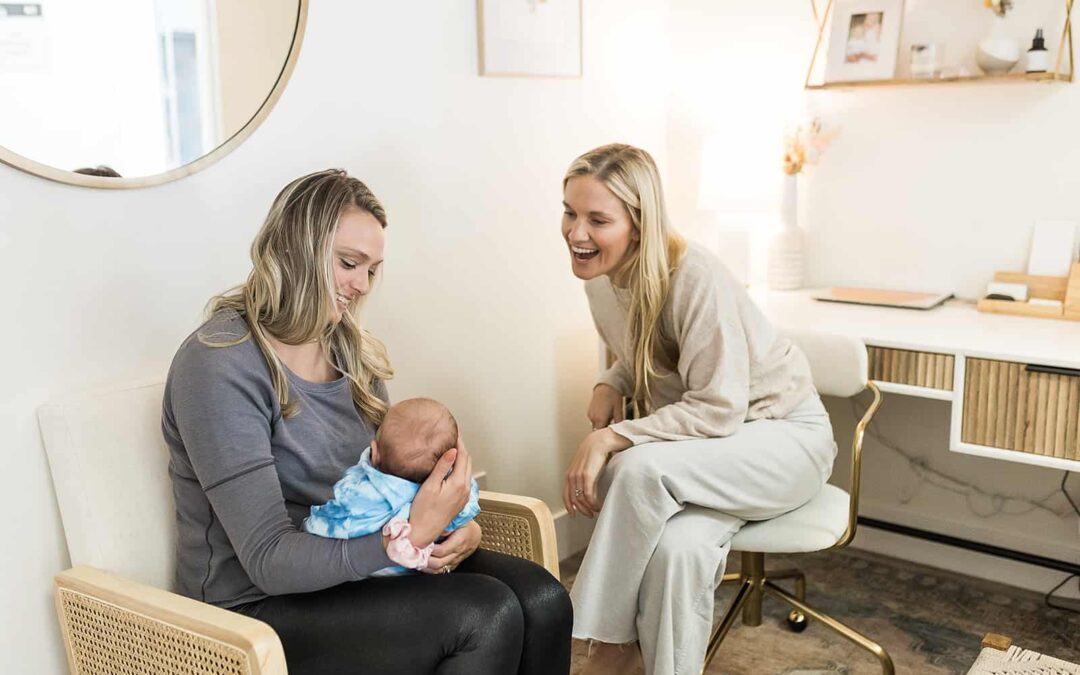 A mom sitting down holding her newborn as another mom smiles at the newborn
