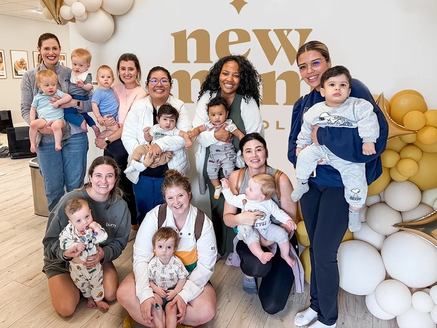 Mothers and Babies Gathering at New Mom School Group of smiling mothers holding babies at New Mom School in front of gold balloon display.