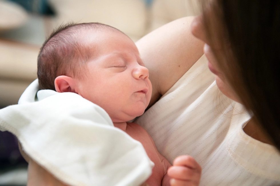 Close-up of a mother gently holding her sleeping newborn wrapped in a white blanket, capturing a quiet and tender moment of early motherhood.