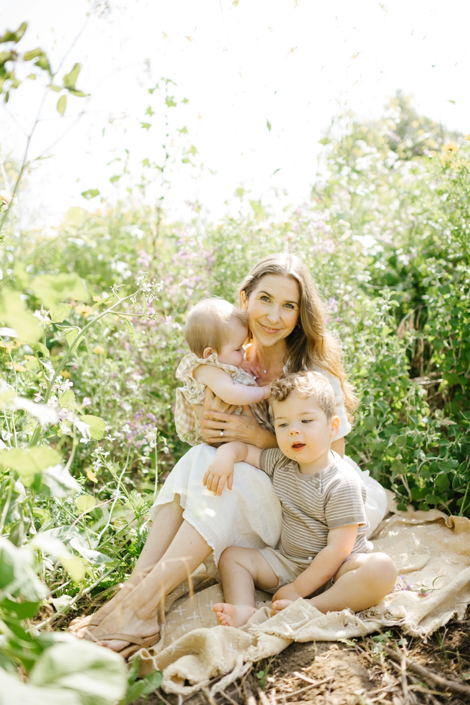 Stephanie Marker, Larchmont Village Location Owner of New Mom School, sitting in a sunlit garden holding her baby while her toddler sits beside her on a blanket.