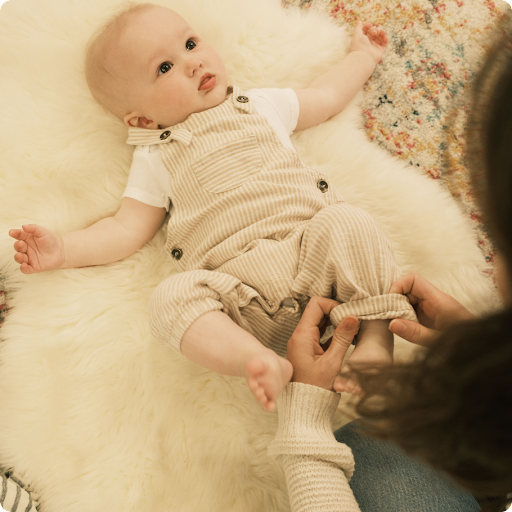 Baby boy in beige overalls lying on a soft rug while a mother adjusts his clothing.